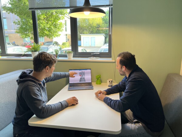 Two people sit at a table, looking at a laptop. One points at a digital 3D model on the screen.