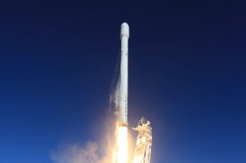 A rocket is launching into a clear blue sky with bright flames and smoke emerging from its engines.