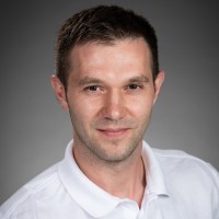 A person with short hair wearing a white polo shirt against a gray background.